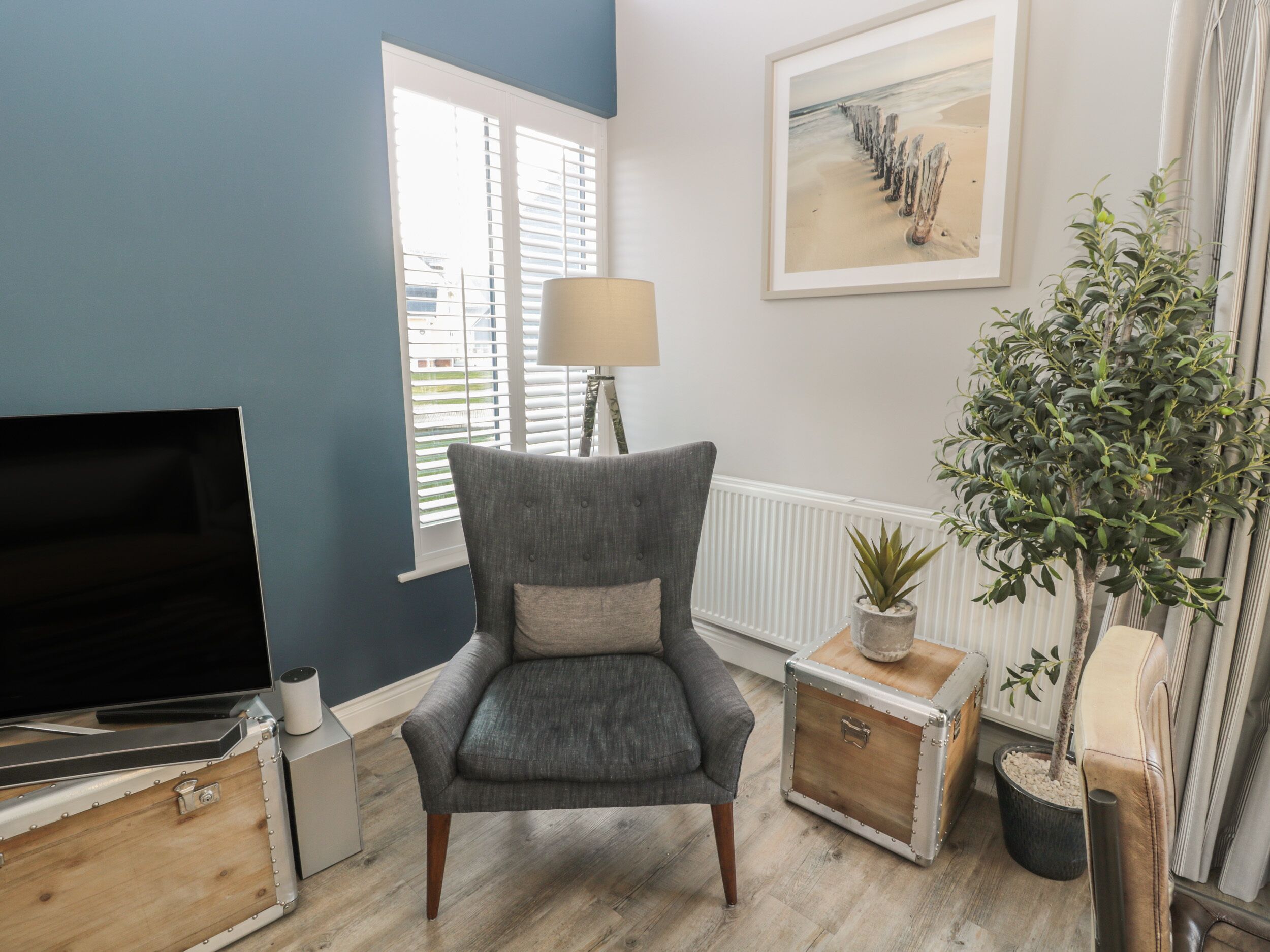 Una sala de estar con un sillón gris, un televisor, una lámpara, un cuadro enmarcado y una planta en maceta.