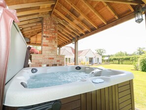Un bain à remous sous une pergola en bois, avec une cheminée en briques, entouré d’une pelouse et d’une maison en arrière-plan.