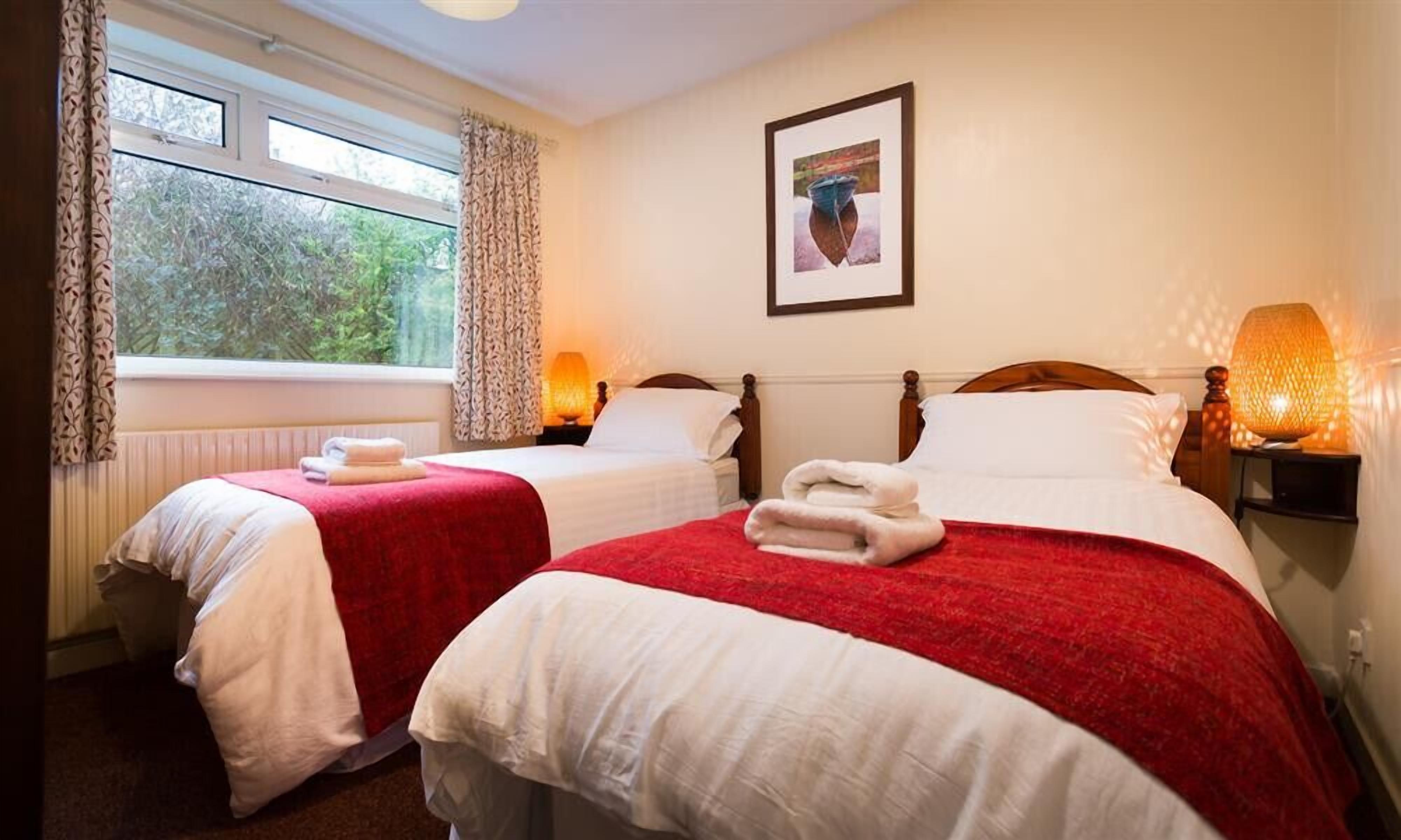 Una camera d'albergo con due letti, ognuno con lenzuola bianche e coperte rosse, una finestra con tende, un quadro appeso al muro e una lampada da comodino.