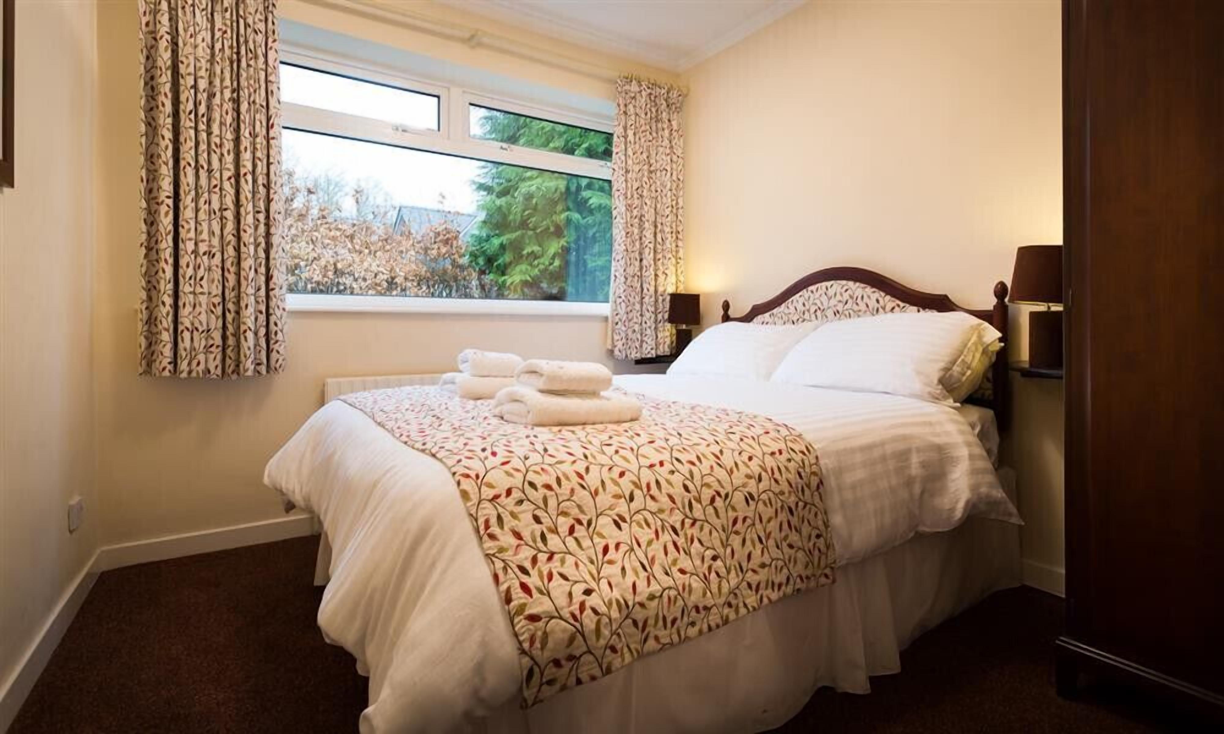 Una cama bien tendida con una colcha estampada, una ventana con cortinas y una lámpara de noche.