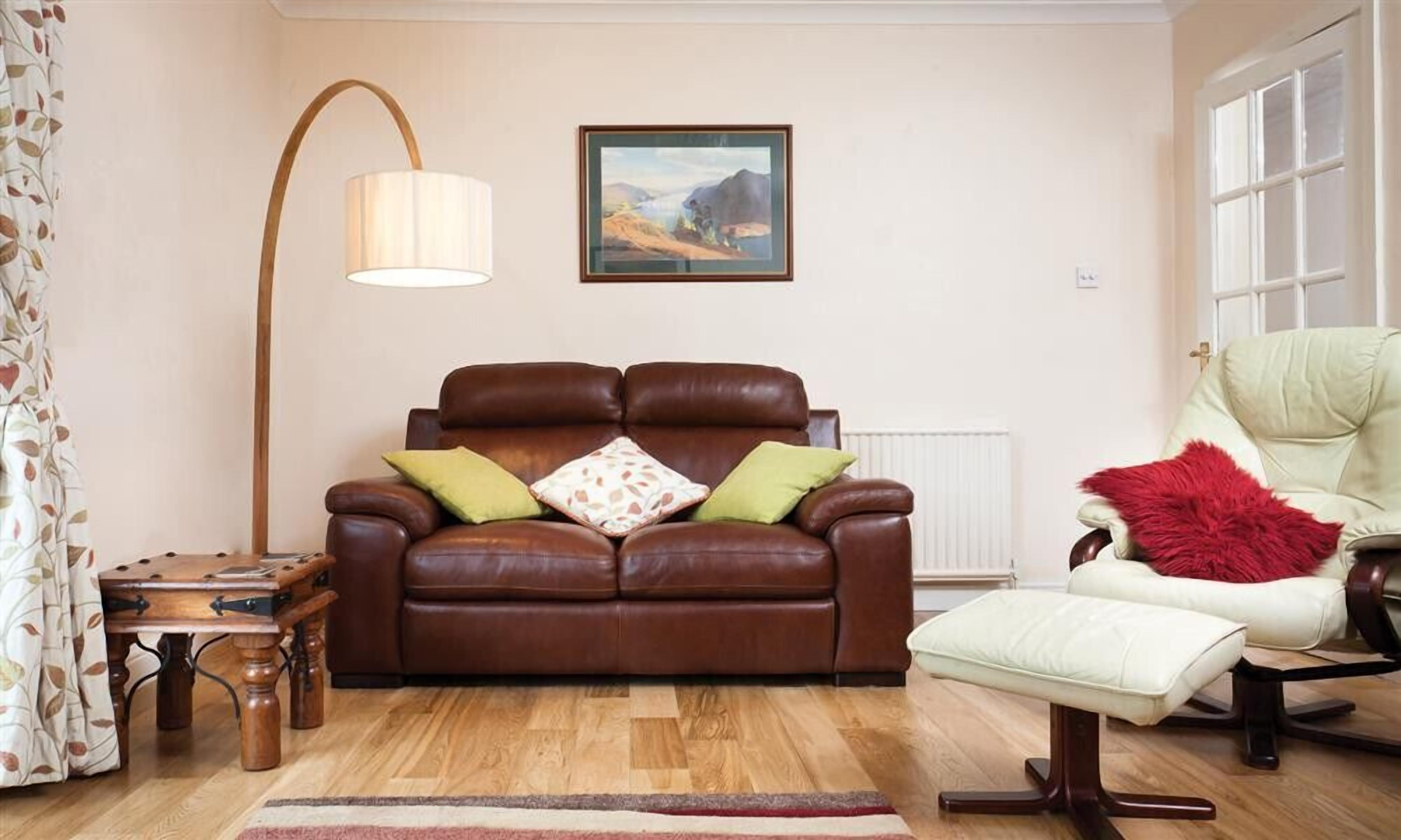 A living room with a brown leather sofa, a white armchair, a wooden side table, a floor lamp, and a painting on the wall.