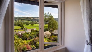A window with a view of a rural landscape, including fields, trees, and a thatched-roof building. - Dormouse Cottage (Coniston)