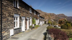 A row of stone buildings with white window frames, a paved road, and mountains in the background. - Dormouse Cottage (Coniston)