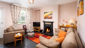 A cozy living room with a fireplace, a sofa, a television, and a window with curtains. - Dormouse Cottage (Coniston)