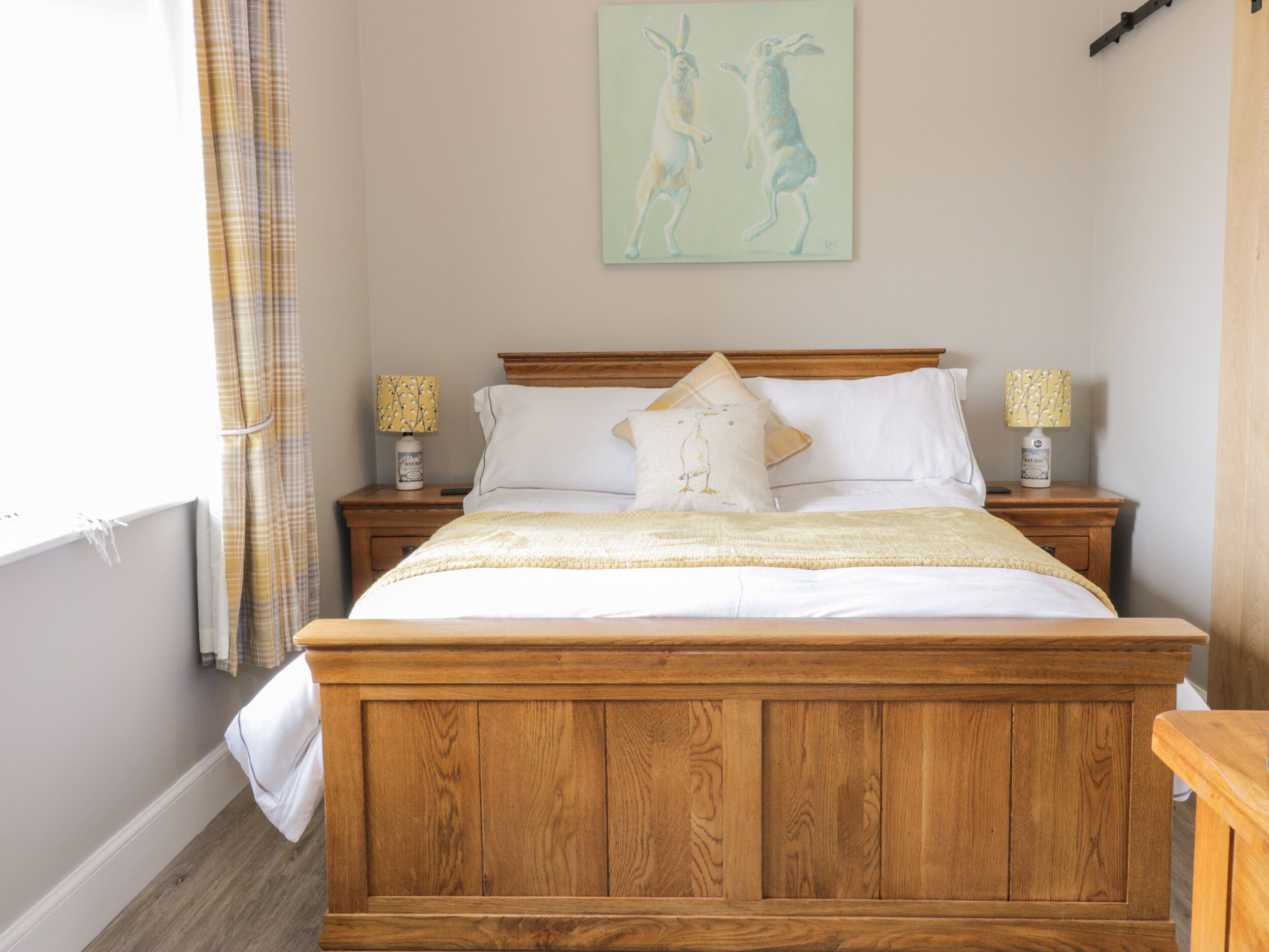 A wooden bed with white bedding, two bedside tables with lamps, and a painting of rabbits on the wall.