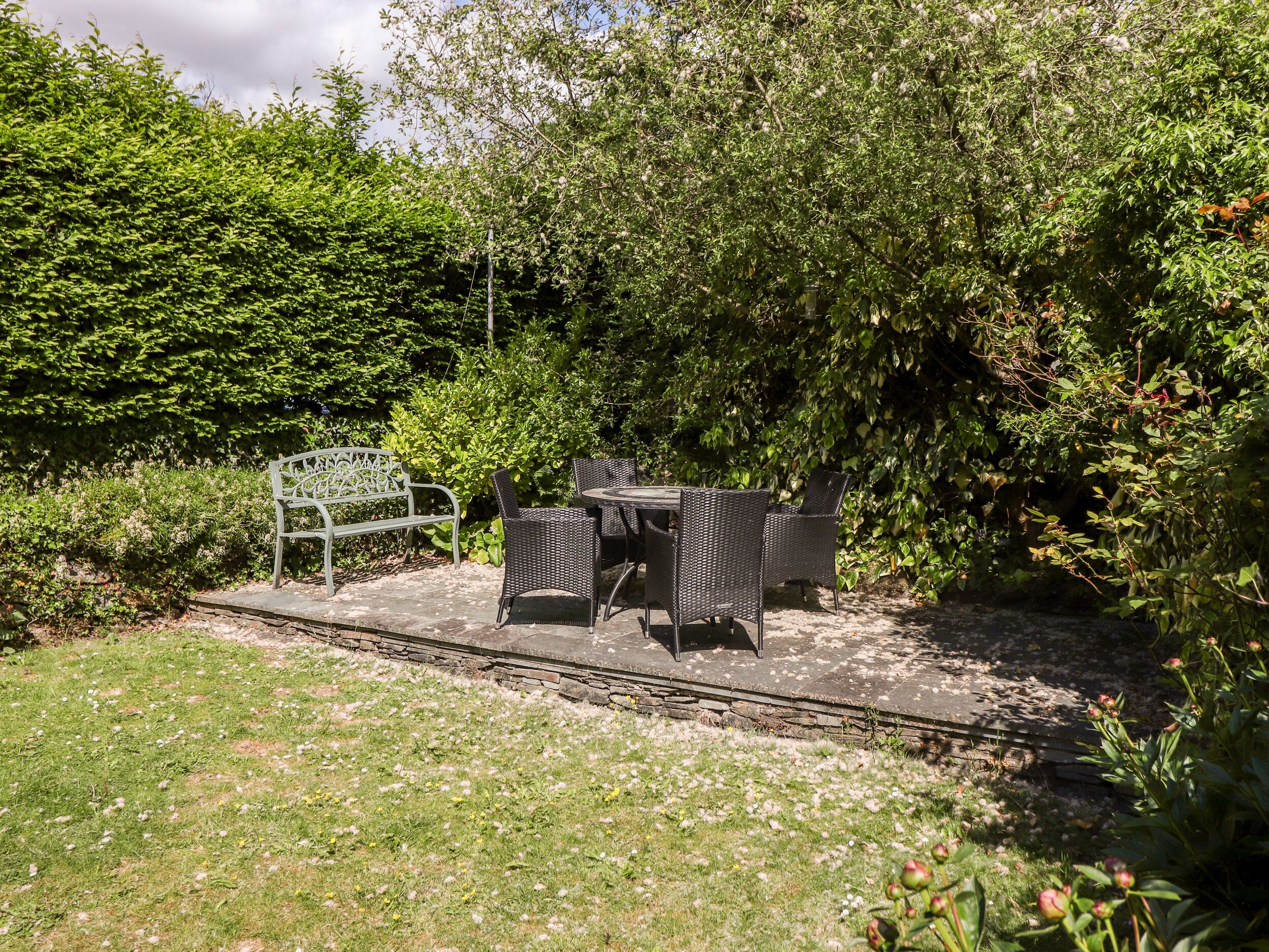A garden patio with a bench and wicker chairs, surrounded by dense foliage.