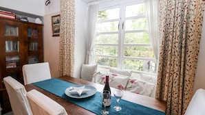 Un coin repas avec une table en bois, des chaises, un chemin de table bleu, une bouteille de vin et des verres à vin. Il y a une fenêtre avec des rideaux et une vue sur la verdure à l’extérieur.