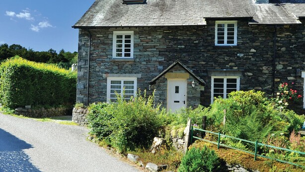 Une maison en pierre avec une porte centrale, deux fenêtres à l’étage supérieur et une cheminée.