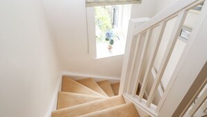 Un escalier dont les marches sont recouvertes d’une moquette beige et dont les rampes sont blanches, menant à un palier agrémenté d’une fenêtre ornée d’une lambrequin à motifs floraux.
