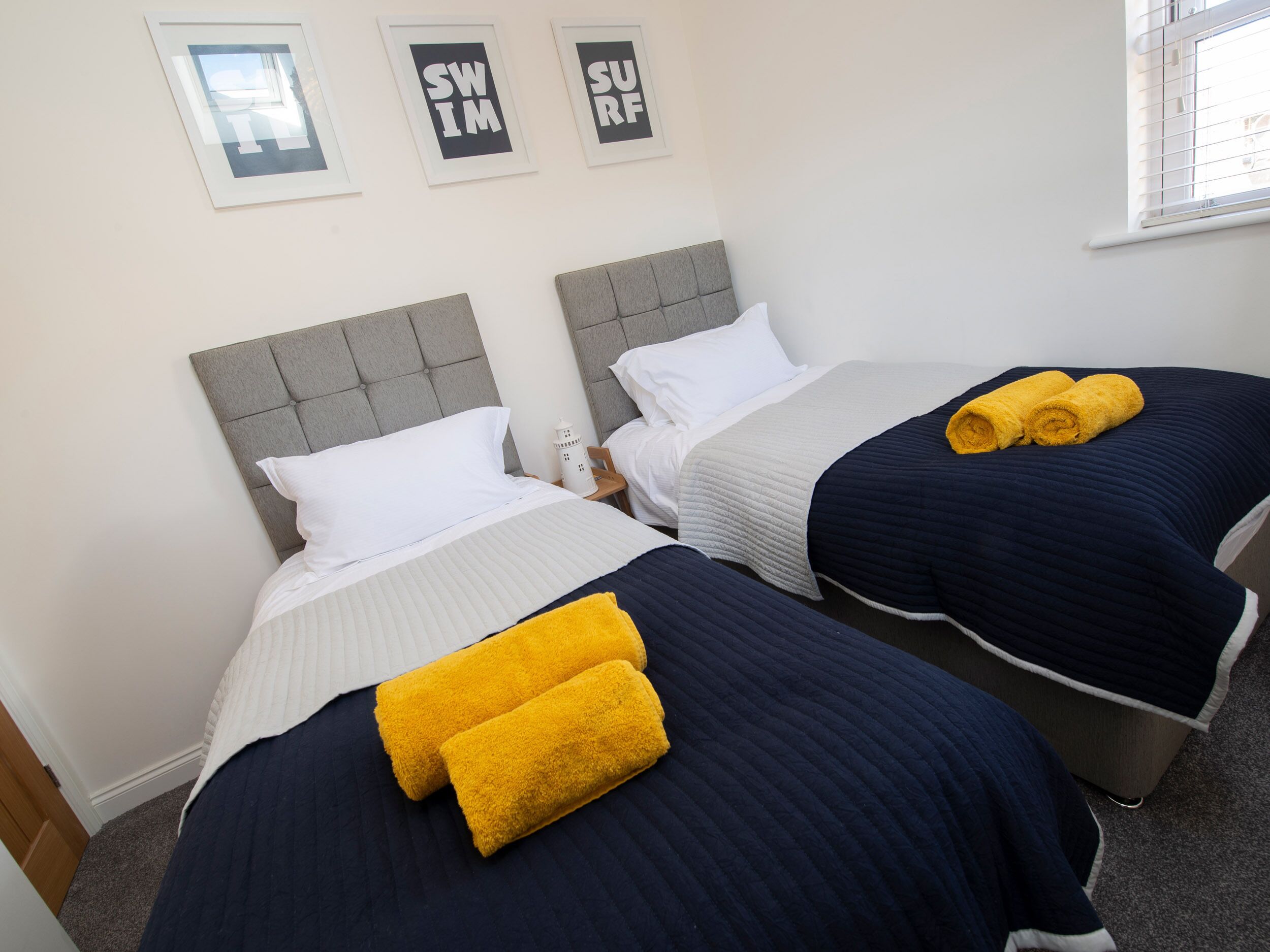 Une chambre d’hôtel avec deux lits, chacun doté d’une tête de lit, de draps blancs et d’une couverture bleu foncé. Les murs sont ornés de tableaux encadrés et il y a une fenêtre avec des stores.