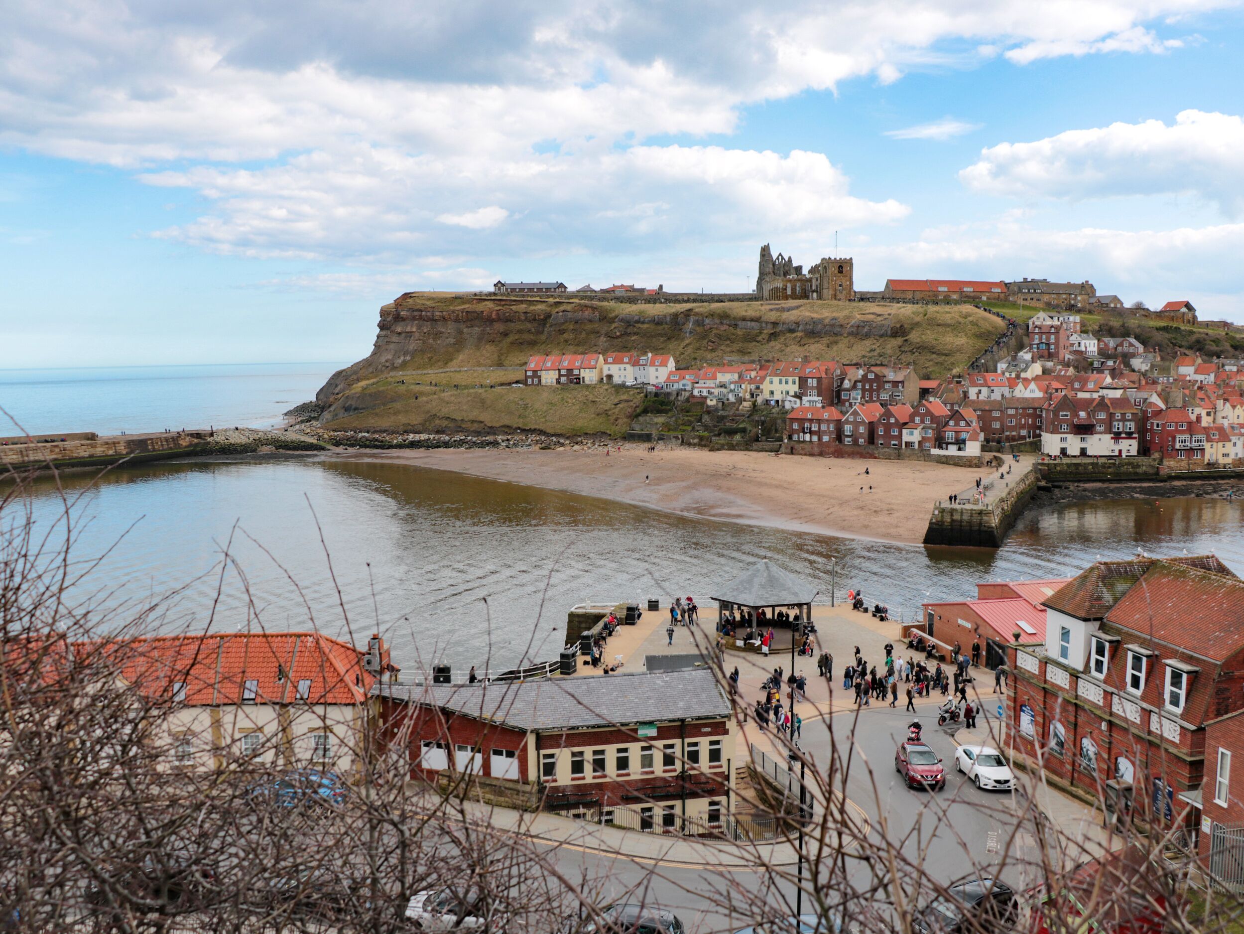 A coastal town with a prominent castle on a cliff, a sandy beach, and a bustling town center with buildings and a road.