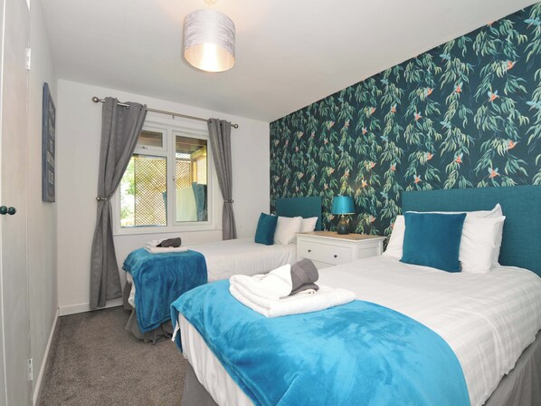 A hotel room with two beds, a window with curtains, a nightstand with a lamp, and a wall with a leafy pattern.