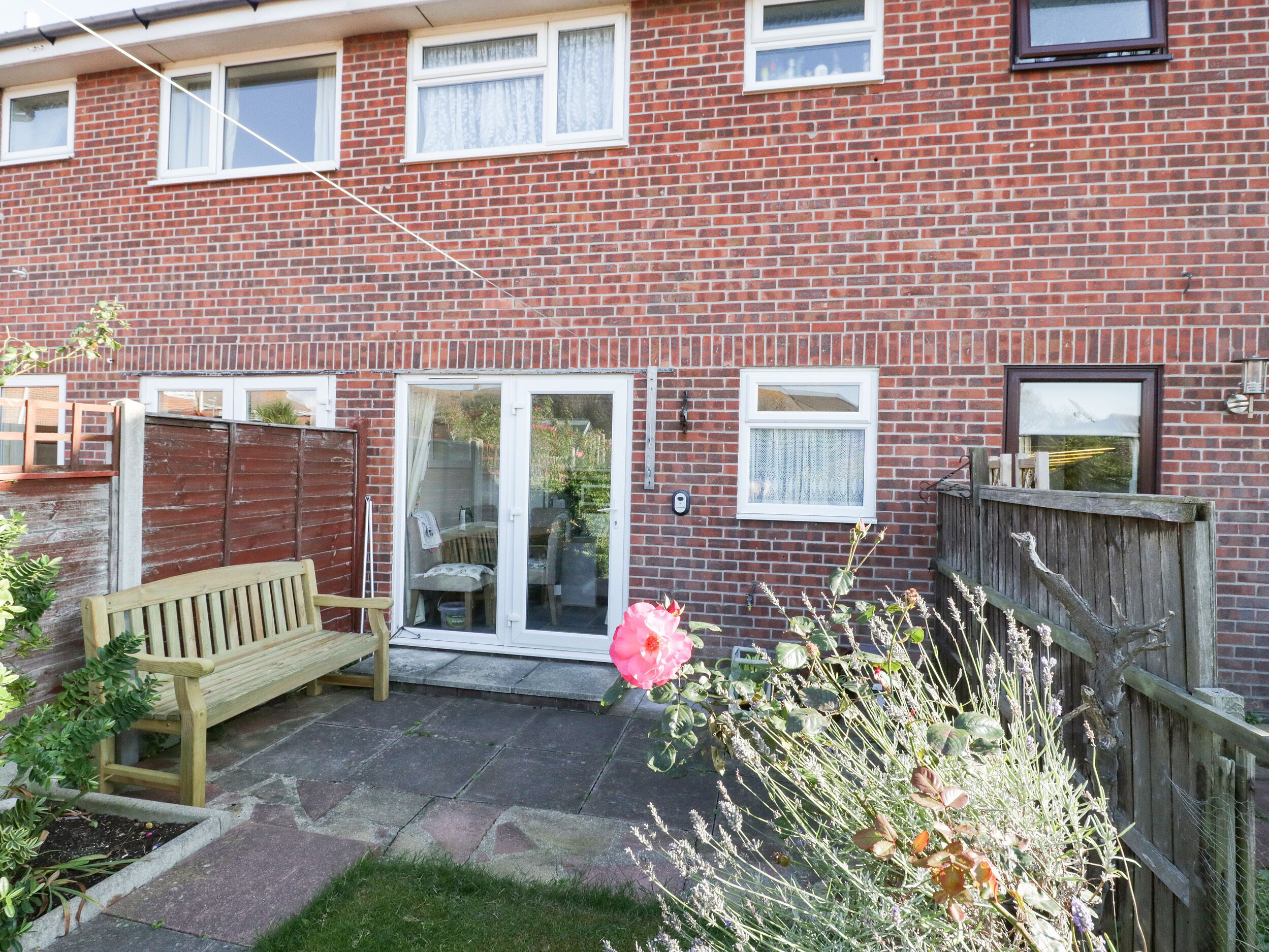 A brick house with a wooden bench, a sliding glass door, and a garden with plants and a pink flower.