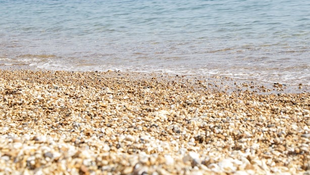 Una spiaggia di ciottoli con un mare calmo e colline in lontananza.
