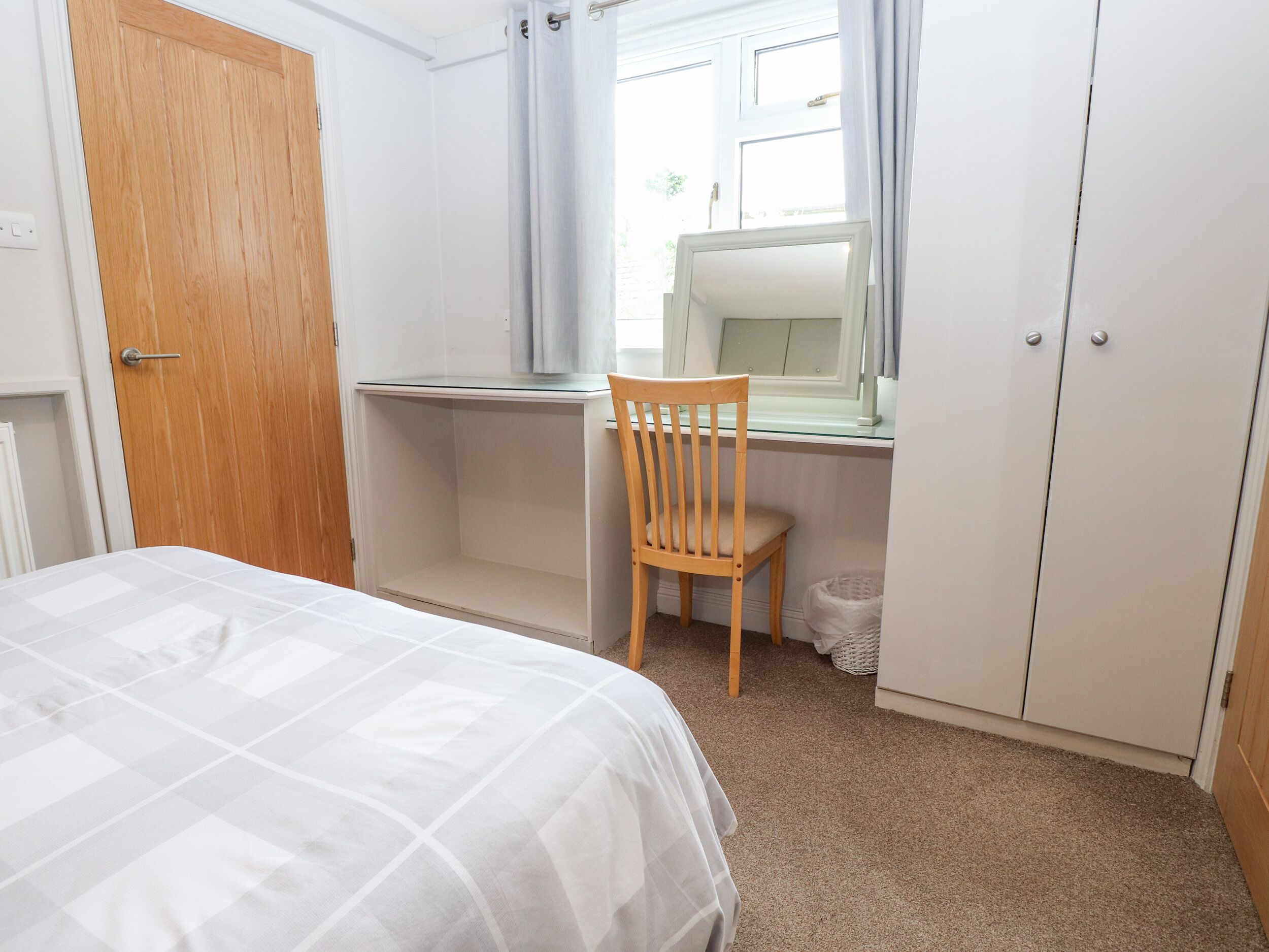 A bedroom with a bed, a wooden chair, a desk with a mirror, and a wardrobe.