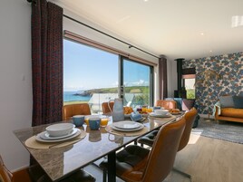 Un comedor con una mesa lista para el desayuno, ventanas amplias con vista al mar, y una sala de estar con sofá y papel tapiz estampado.