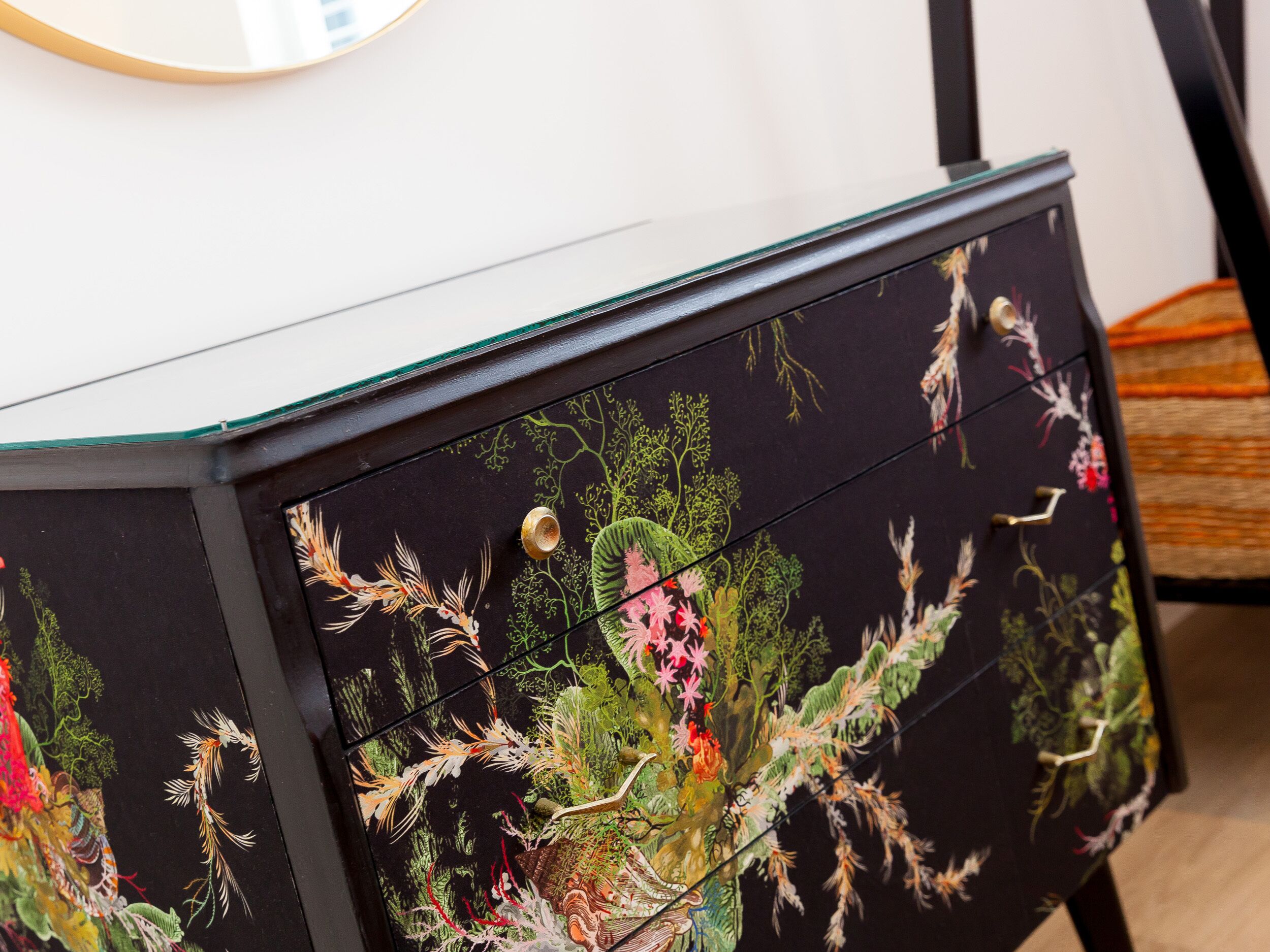 Eine schwarze Kommode mit floralen und blattförmigen Mustern, darüber ein runder Spiegel und ein Holzboden.
