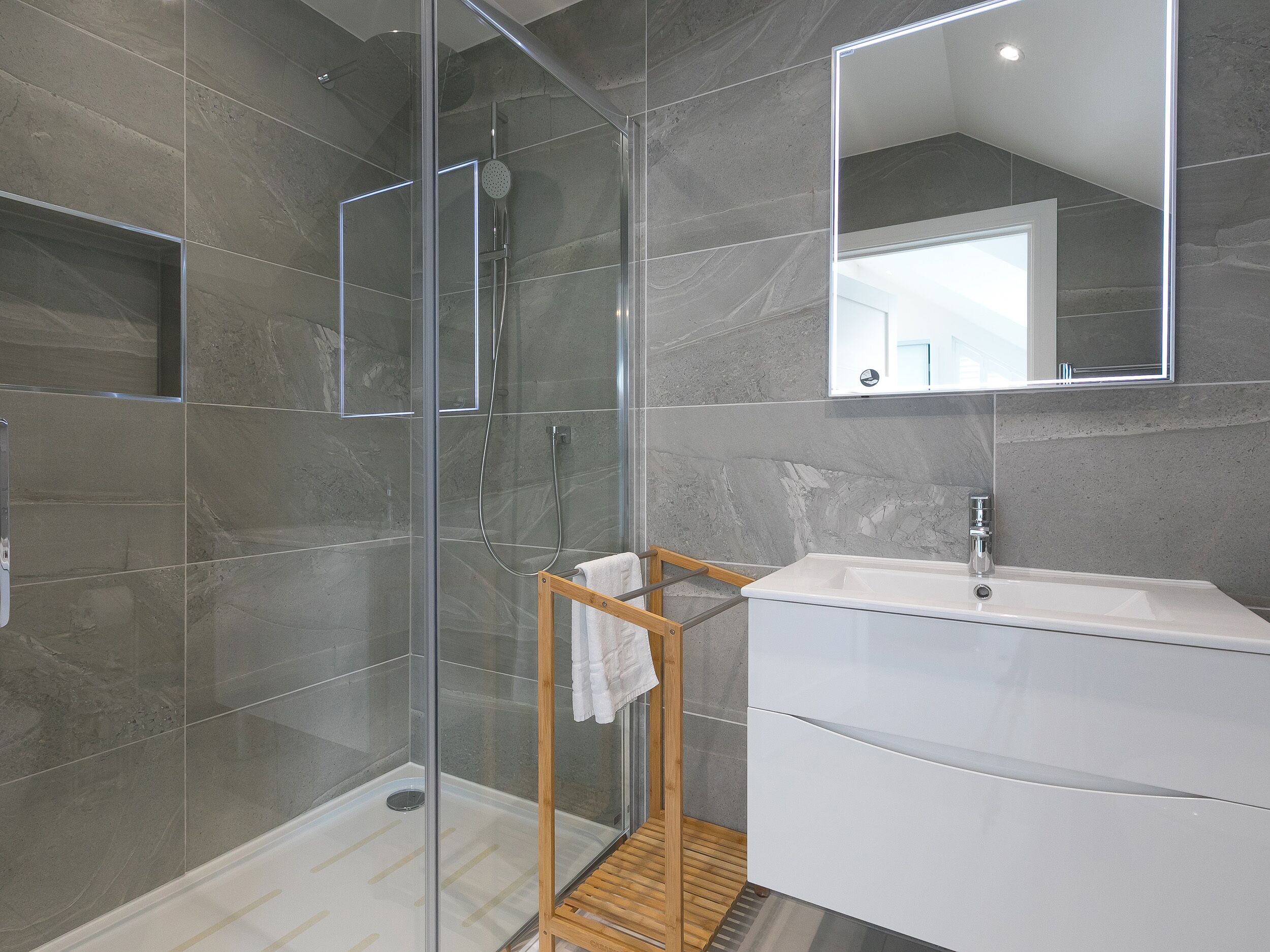 A modern bathroom with a glass-enclosed shower, a wooden towel rack, and a white sink.