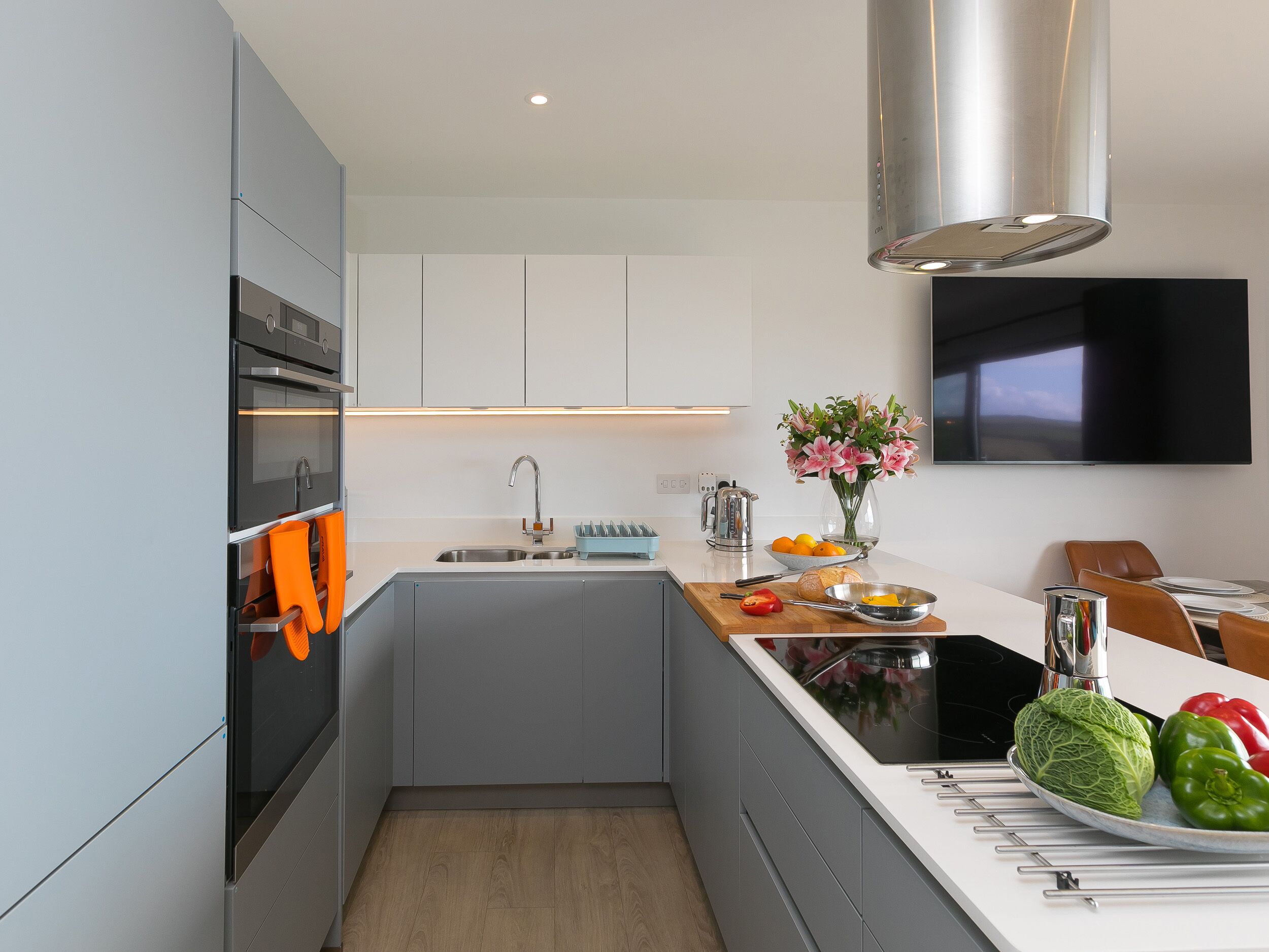 Une cuisine moderne avec des armoires grises, une hotte de cuisinière en acier inoxydable, un four encastré, un évier et un coin repas avec un téléviseur placé au-dessus du comptoir.