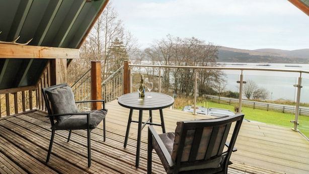 A wooden deck with two chairs and a table, overlooking a lake and hills. - The Apartment (Fort William)