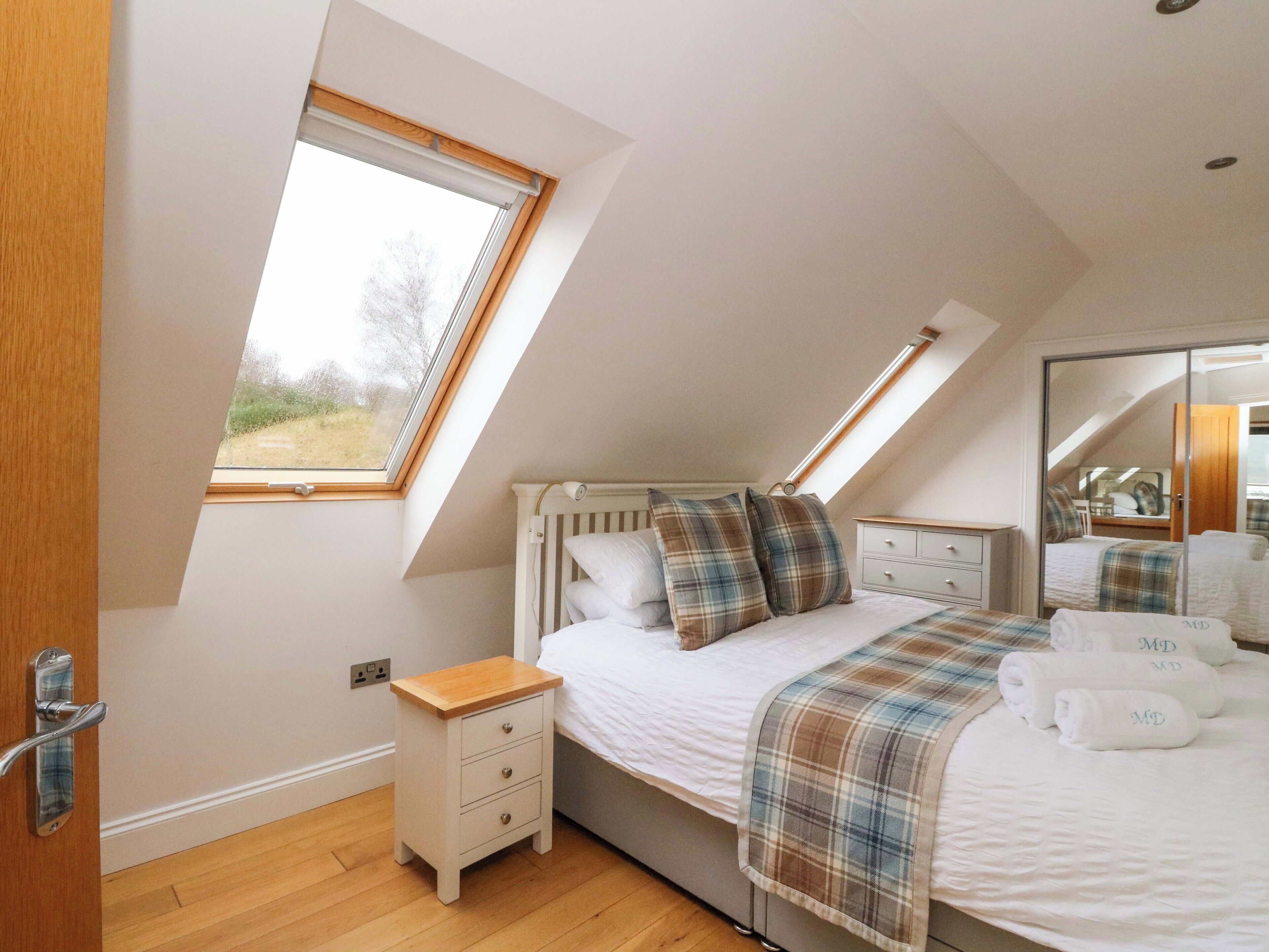 A bedroom with a sloped ceiling, a skylight, a bed with a plaid blanket, a bedside table, and a mirror.