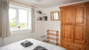 Un dormitorio con un armario de madera grande, una cama con colcha estampada, una ventana con cortinas, un radiador y un estante pequeño con libros y un jarrón.