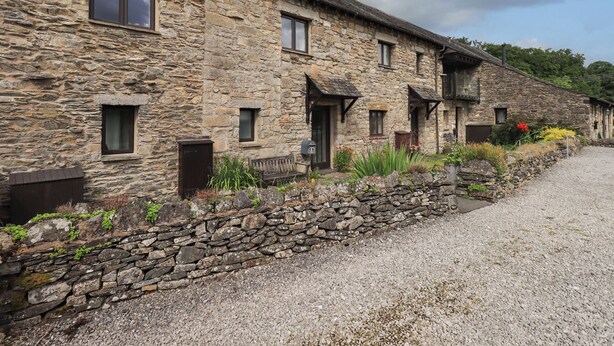 A stone building with multiple windows and doors, a gravel driveway, and a stone wall with plants. - Tullythwaite Garth (Kendal)