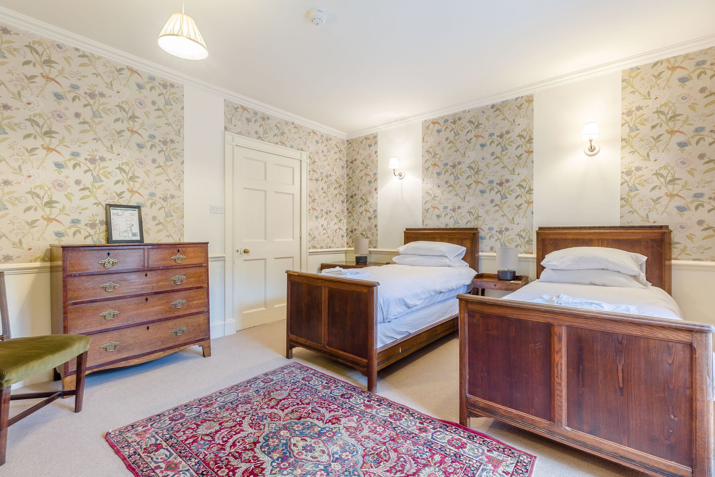 Un dormitorio con dos camas de madera, un aparador, una silla y una alfombra estampada.