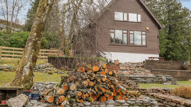En rustik hytte med stensokkel og trætag, omgivet af en stenvæg og en bunke brænde.