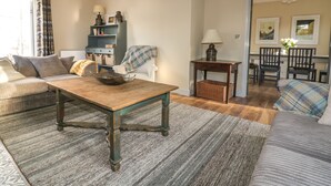 Un salon chaleureux avec un canapé, une table basse, une table d’appoint et une lampe. On aperçoit une bibliothèque et un fauteuil au fond de la pièce.