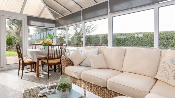 A sunroom with a large glass door, a cushioned sofa, a dining table with chairs, and a wicker coffee table with a magazine and a vase. - Bryngwyn - Trearddur Bay (Holyhead)