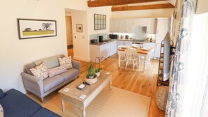 A spacious living area with a sofa, coffee table, and dining table set for four. A kitchen with white cabinets and a wooden ceiling is visible in the background.