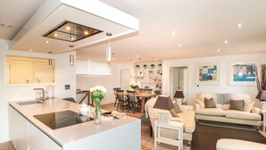 A modern kitchen with a central island, a dining area with a table and chairs, a sofa, and a bookshelf. - 2 Sails (Dartmouth)