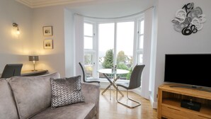 A living room with a grey sofa, a TV, a wooden coffee table, and a dining area with chairs and a glass table. - Tethera (Windermere)