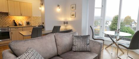 A modern living room with a grey sofa, two patterned cushions, a dining table with chairs, a kitchen area with wooden cabinets, and a large window offering a view of the outside.