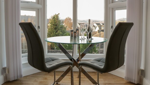 A modern dining area with a glass table and two chairs, large windows with curtains, and a view of the outdoors. - Tethera (Windermere)