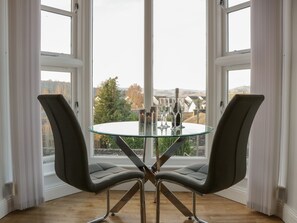 Un espace repas moderne avec une table en verre et deux chaises, de grandes fenêtres avec des rideaux et une vue sur l’extérieur.