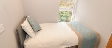 A small, neatly made bed with a white and blue bedspread, a single pillow, and a window with a view of greenery outside.