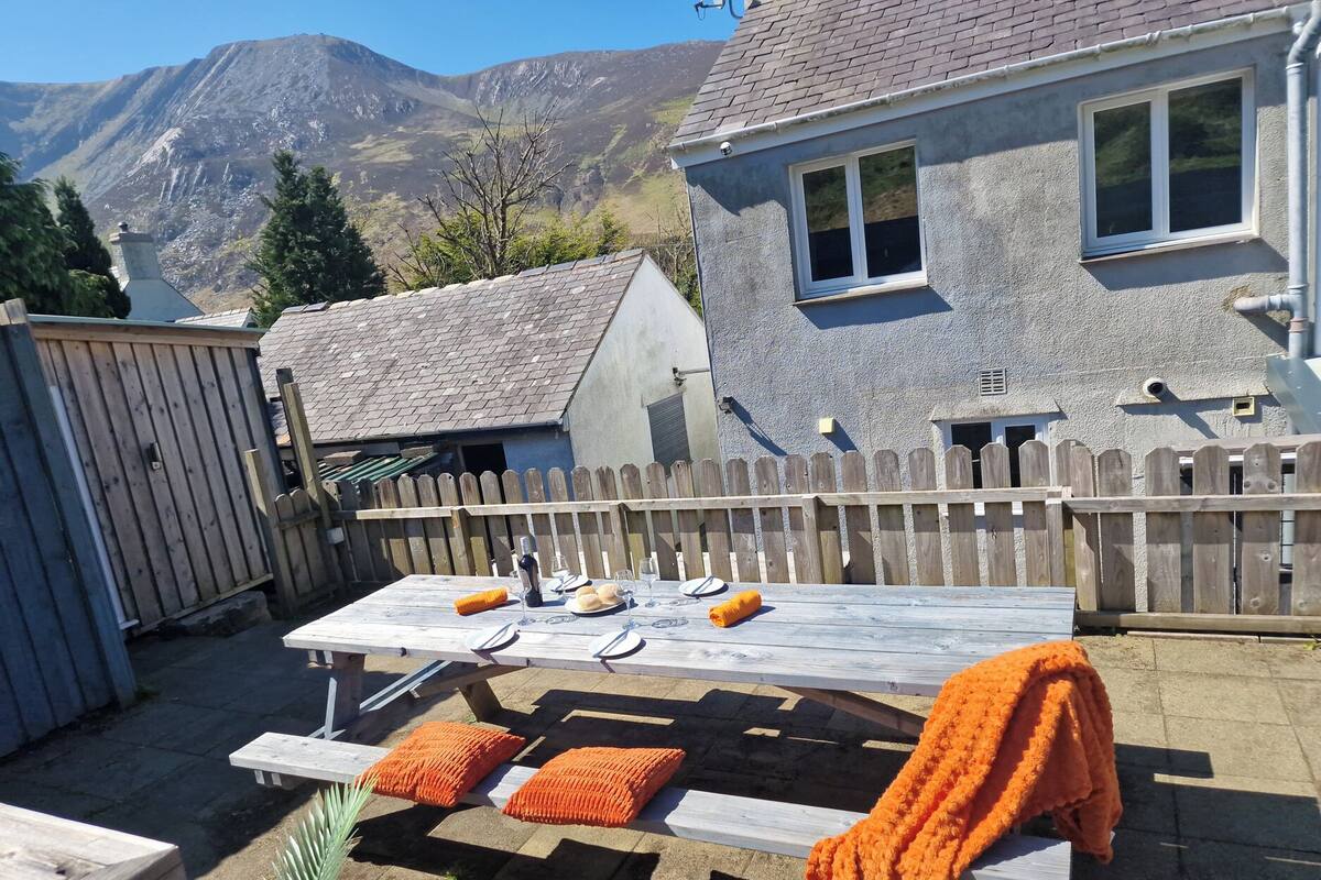 Ett träbord placerat utomhus med en bergskedja i bakgrunden, ett trästaket och ett hus med en grå fasad.