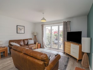 A modern living room with a brown leather sofa, a television on a wooden cabinet, and a large sliding glass door leading to an outdoor patio. - Buck Place (Filey)