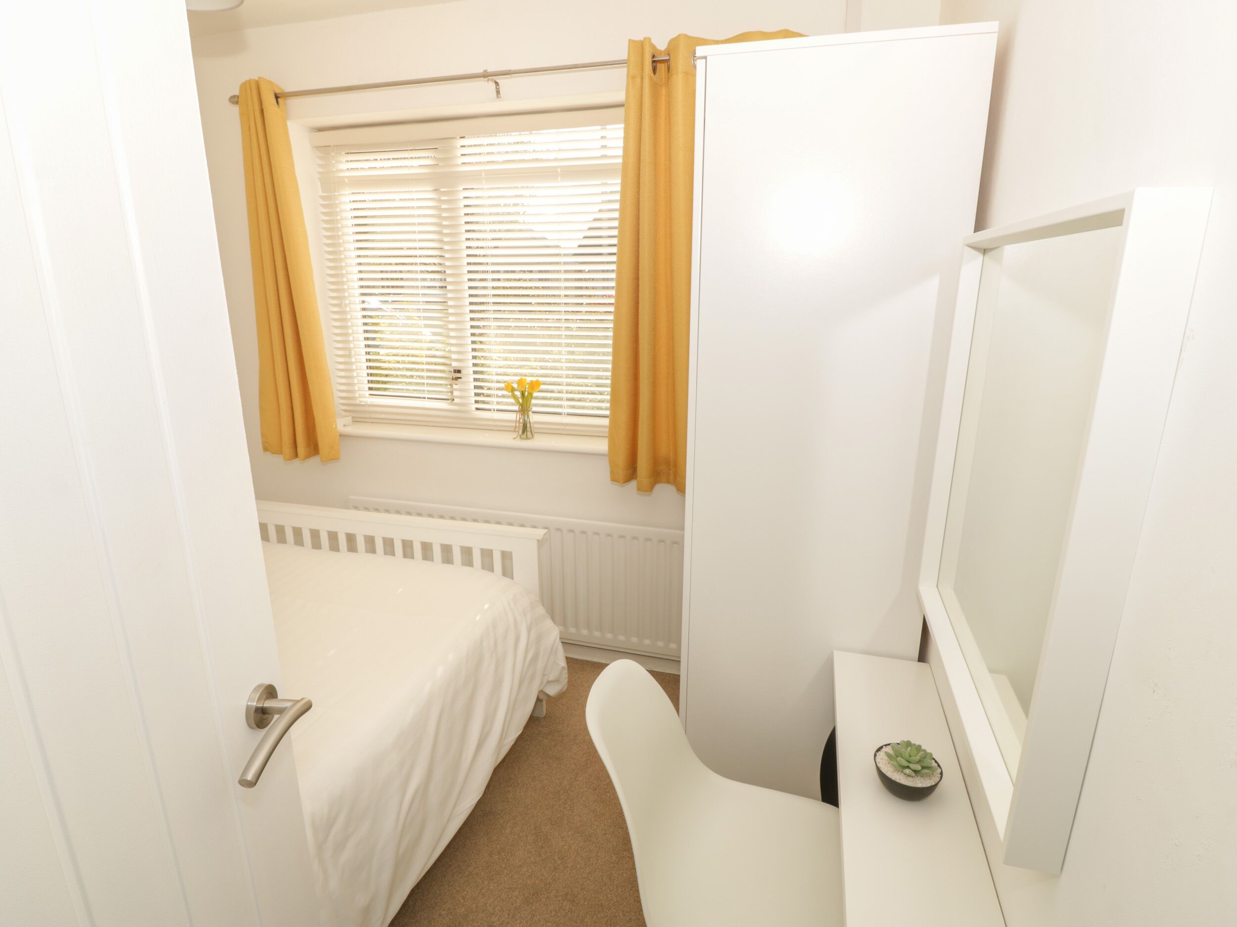 Un dormitorio pequeño y ordenado con una cama individual, una mesita de noche con una planta y una ventana con cortinas.