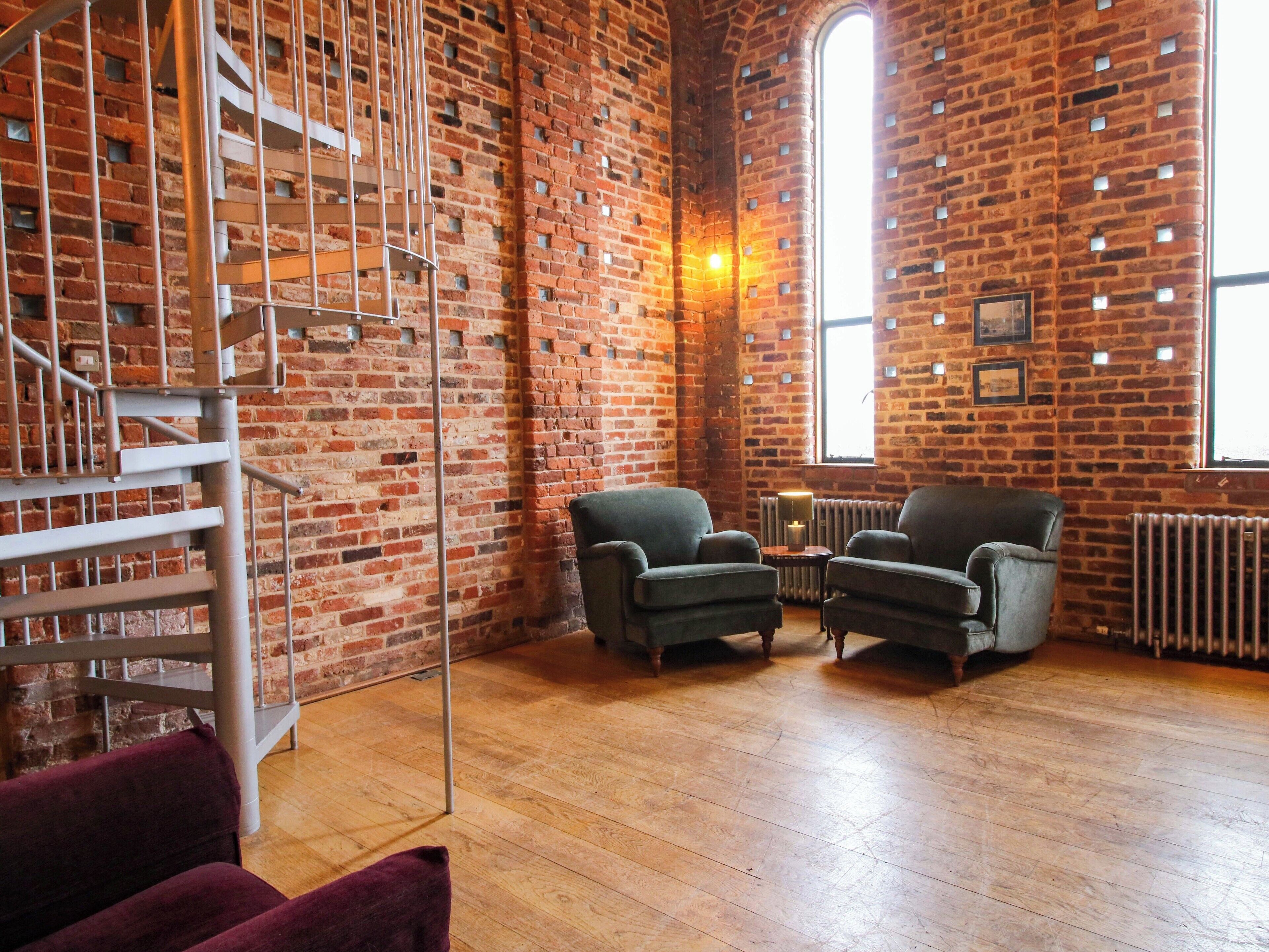 A room with exposed brick walls, a spiral staircase, two armchairs, a small table with a lamp, and a radiator.