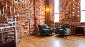 A room with exposed brick walls, a spiral staircase, two armchairs, a small table with a lamp, and a radiator.