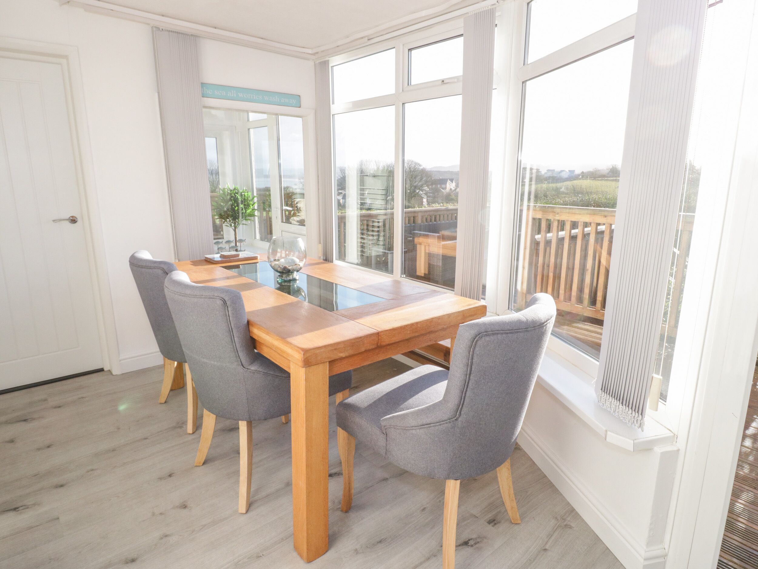 A dining area with a wooden table and grey upholstered chairs, large windows, and a view of the outdoors.
