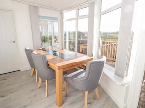 Un coin repas avec une table en bois et quatre chaises grises, de grandes fenêtres donnant sur l’extérieur et une porte située à gauche.