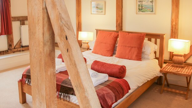 Un lit à ossature en bois, agrémenté de coussins rouges et d’une couverture, deux lampes de chevet et une petite table en bois.