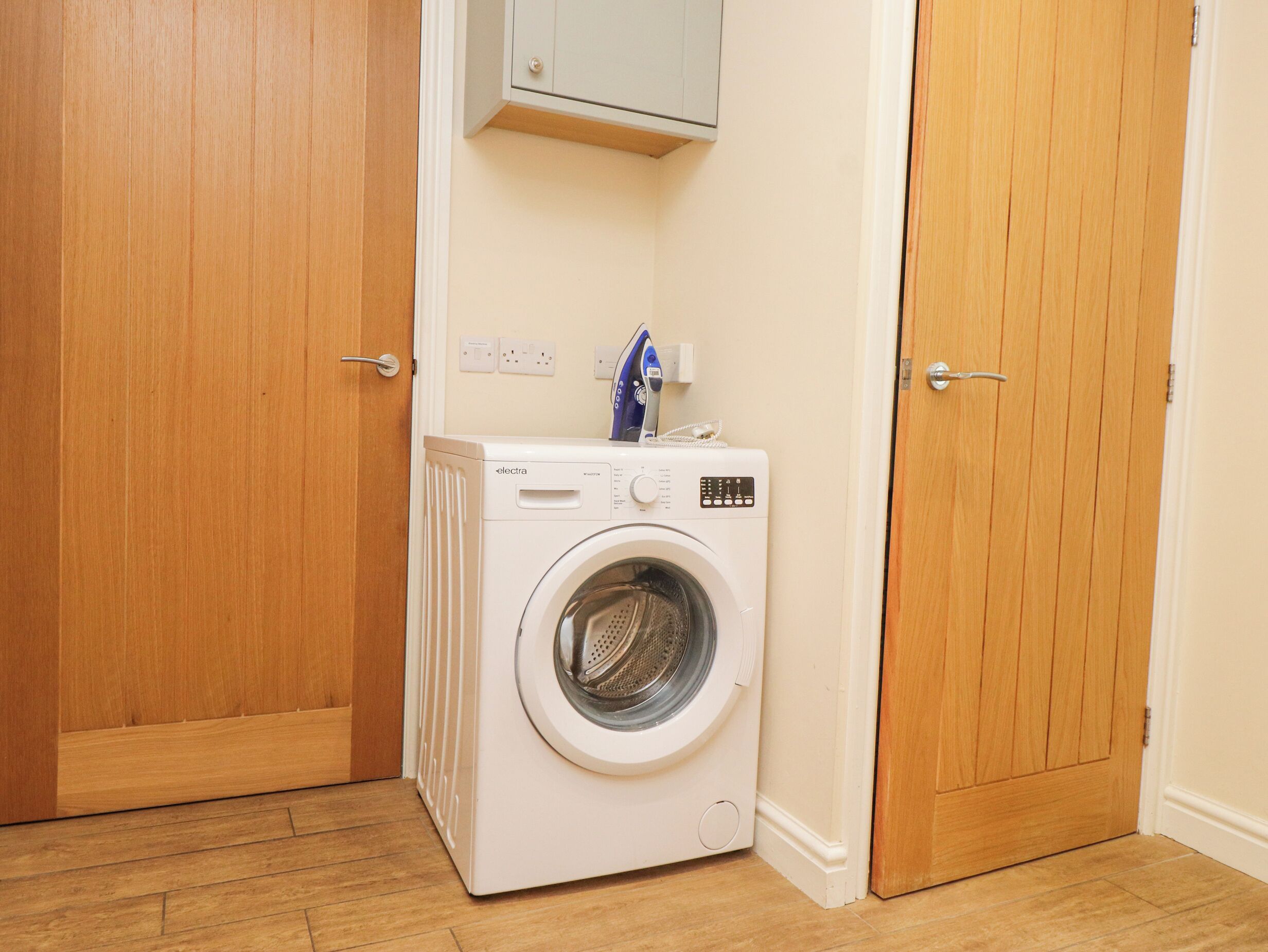 A compact, white washing machine in a small laundry room with wooden doors and a light-colored floor.