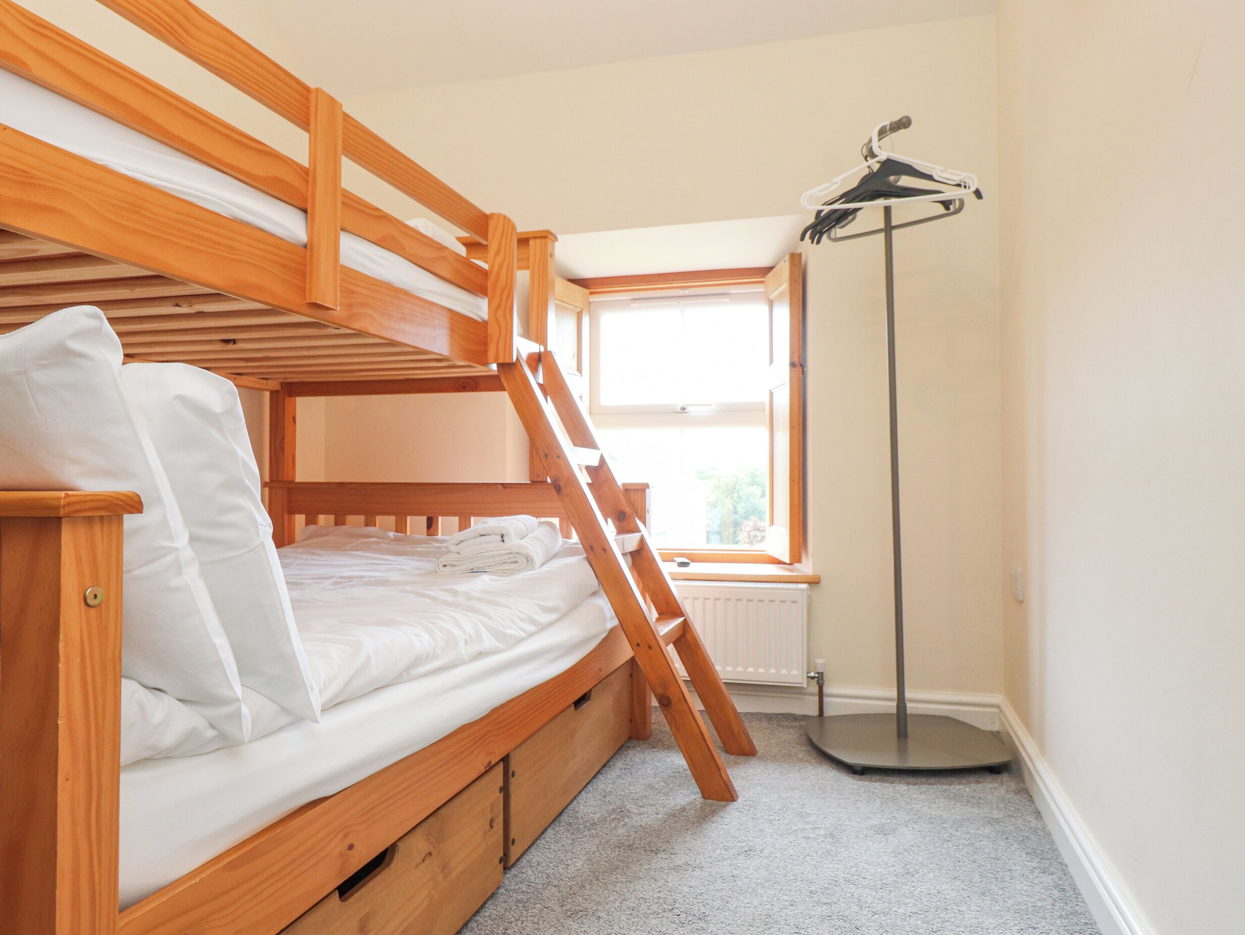 A bunk bed with a wooden ladder, a radiator, and a window with a view of greenery.