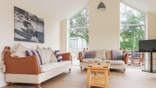 A spacious living room with two sofas, a coffee table, and a TV. Large windows provide a view of the outdoors.