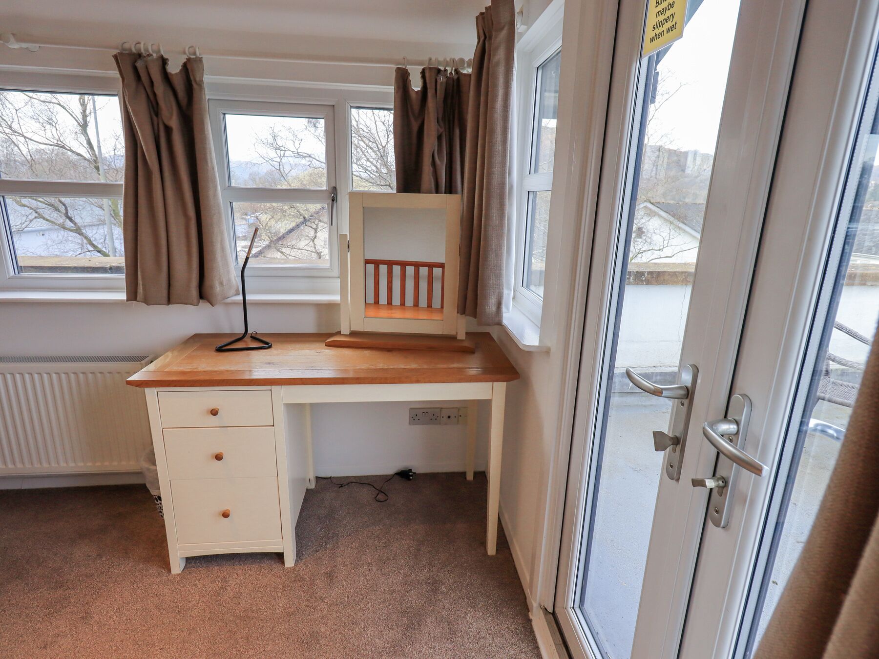 A small, tidy room with a wooden desk, a radiator, and a window with curtains.